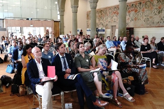 Teilnehmende einer Konferenz sitzen in einem großen Saal mit Marmorsäulen und sind während eines Vortrags oder Meetings zu sehen. Mehrere Personen sitzen in Reihen auf Stühlen und schauen nach vorn; einige halten Unterlagen oder Notizhefte.
