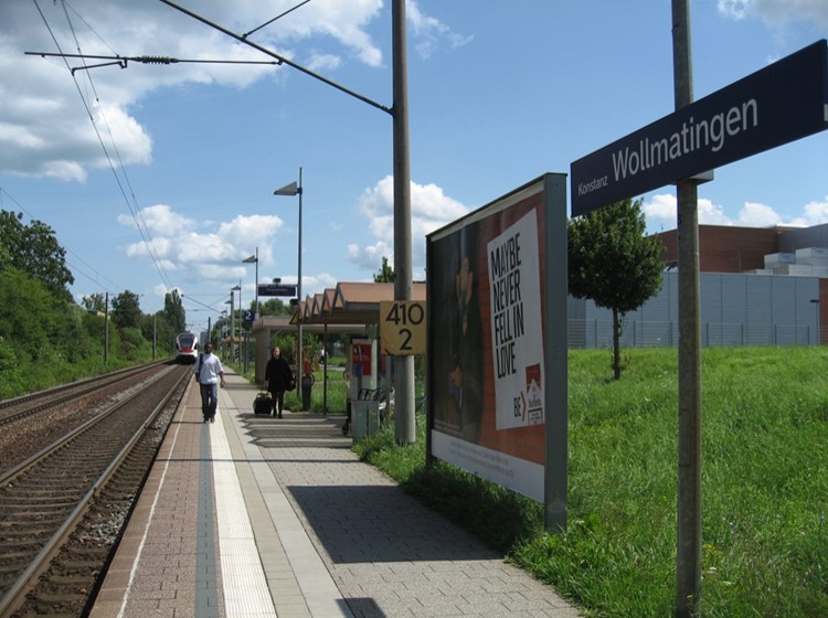 Auf dem Bild zu sehen ist der Wollmatinger Bahnhof. Schienen, Gleisbett, Bahnsteig, viele Schilder und entfernt Menschen.