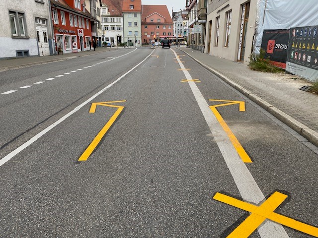 Das Bild zeigt eine Straße in einer Stadt mit frisch aufgebrachten gelben Fahrbahnmarkierungen. Zu sehen sind große Pfeile und Kreuze auf dem Asphalt, die den Verkehr leiten sollen. Entlang der Straße stehen mehrstöckige Gebäude mit Geschäften im Erdgeschoss.