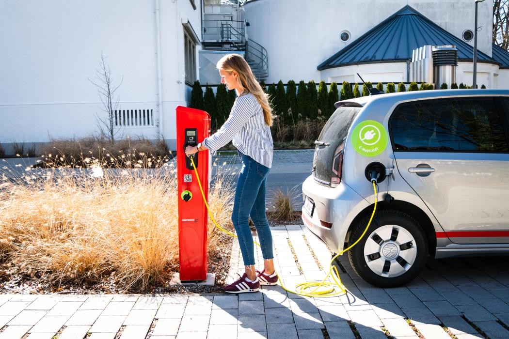 Eine Frau lädt ein kleines silberfarbenes E-Auto an einer roten Stromladesäule