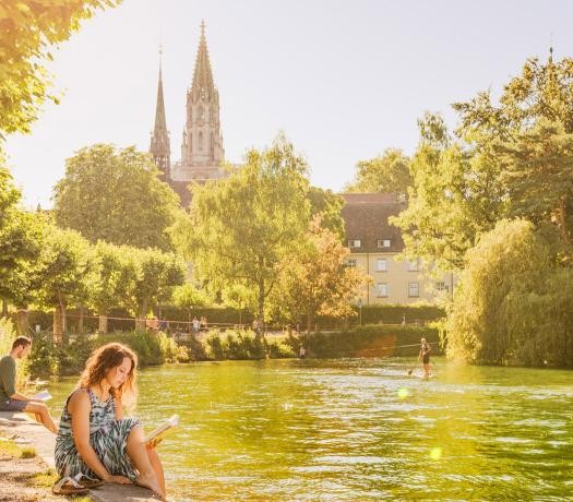 Menschen sitzen am Wasser; drumherum eine parkähnliche Anlage