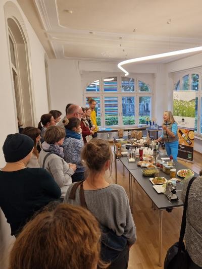 Eine Personengruppe steht vor einem langen Tisch, auf dem Speisen aufgetischt sind, während eine Frau einen Vortrag hält