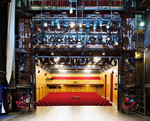 Blick von der Bühne in den Zuschauersaal mit roten Sesseln und holzverkleideten Wänden; über der Bühne zahlreiche Scheinwerfer