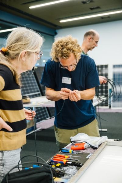 Zwei Personen an einem Tisch, auf dem Werkzeuge liegen; im Hintergrund Solarpaneele