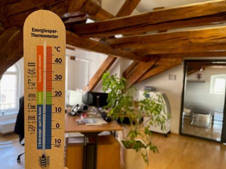 im Vordergrund ein hölzernes Thermometer, das 19 Grad Celsius anzeigt; im Hintergrund ein Büroraum