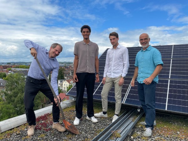 Vier Männer stehen auf einem Dach vor einer Photovoltaik-Anlage. Ein Mann hält eine Schaufel in der Hand.