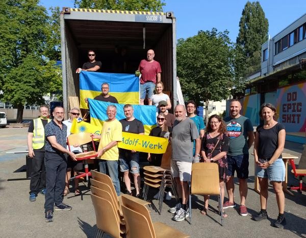 Ein Laster mit offener Verladeklappe. Davor mehrere Menschen, und Stühle. Eine Ukrainefahne wird gehalten.