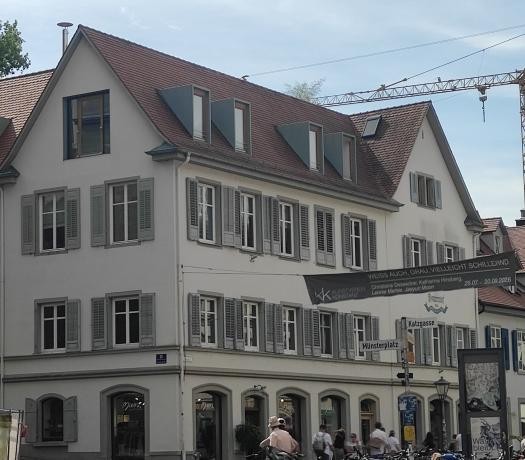 Straßenflucht mit Blick auf ein weißes Eckhaus mit rotem Dach und grünen Fensterläden