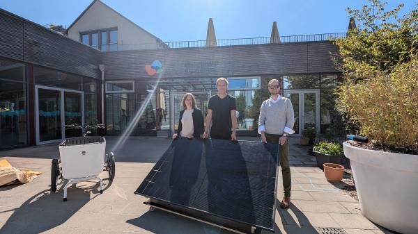drei Personen stehen draußen vor dem Kinderhaus Hl. Dreifaltigkeit. Vor ihnen steht ein Solar-Modul auf einem Anhänger. Daneben ein Lastenrad.