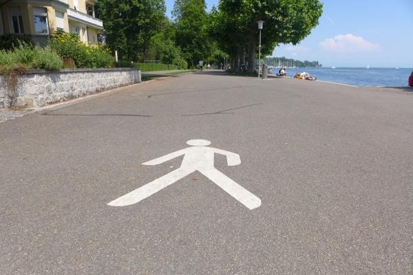 Das Bild zeigt eine breite Uferpromenade mit einem großen weißen Fußgängersymbol auf dem Asphalt. Links sind Blumen, ein Haus und eine niedrige Mauer zu sehen, rechts das Ufer mit Menschen am Wasser und Segelbooten auf dem See. Der Weg ist eben und gut zugänglich für Fußgänger.