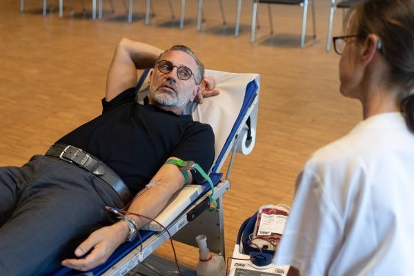 Copyright Stadt Konstanz Auf dem Foto liegt ein Mann auf einer Liege. Er spendet Blut. Sein Arm ist ausgestreckt. An seinem Arm ist eine Nadel. Daran hängt ein Beutel mit Blut. Rechts vorne sitzt eine Frau. Man sieht sie von hinten.