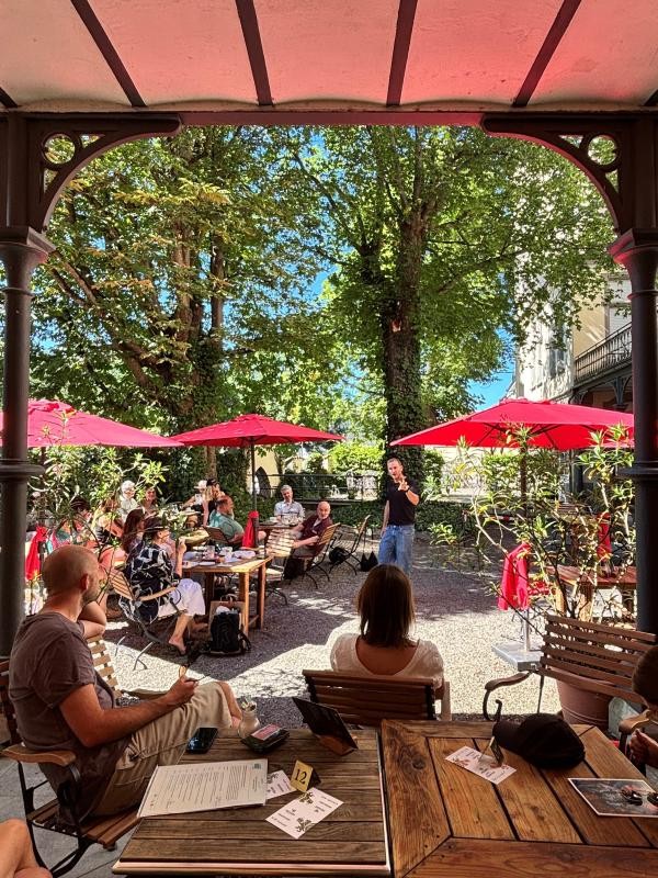 Eine Person spricht vor mehreren Menschen, die unter Schirmen an Tischen in einem Garten eines Cafés sitzen