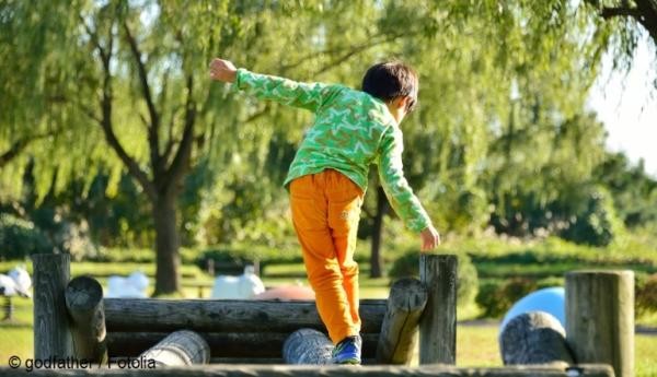Ein Kleinkind in grünem Shirt und orangener Hose balanciert auf einem Geländer. Es ist von hinten zu sehen und streckt die Arme zur Seite aus.