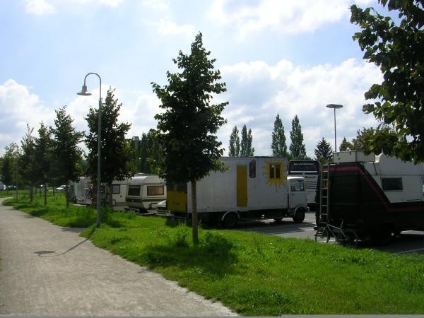 Das Bild zeigt mehrere Wohnwagen und Fahrzeuge, die auf einem Parkplatz entlang einer Baumreihe abgestellt sind. Links daneben verläuft ein Fußweg mit Laternen und einer grünen Wiese. Der Himmel ist leicht bewölkt, die Szene wirkt ruhig und sommerlich.