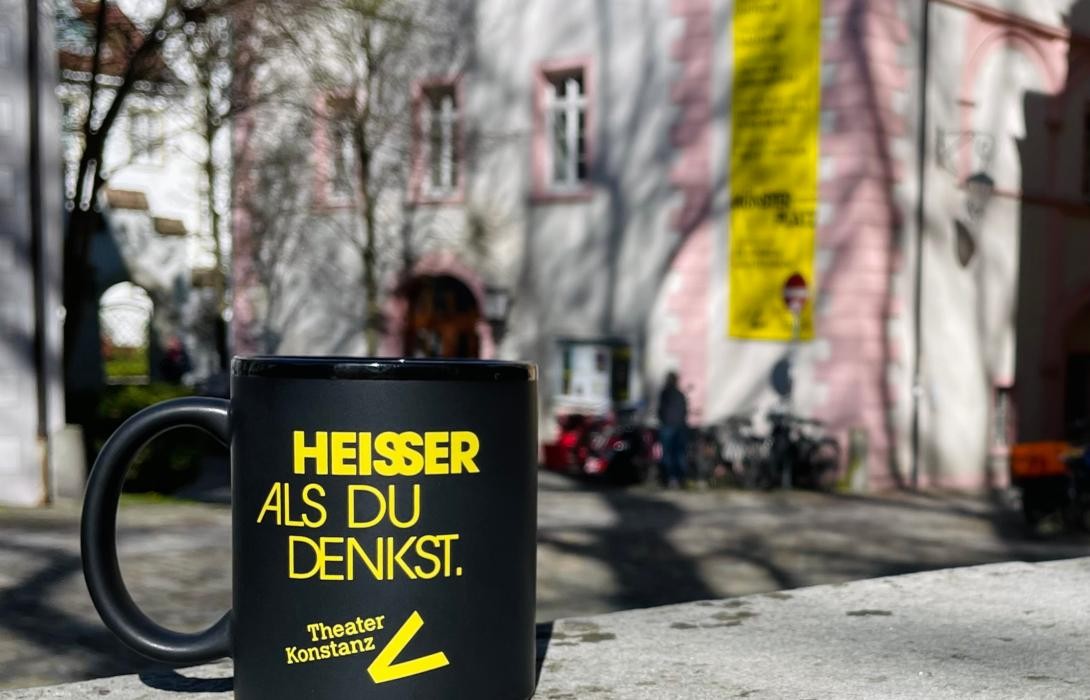Eine schwarze Tasse mit gelber Schrift "Heisser als du denkst", im Hintergrund sieht man das Theatergebäude