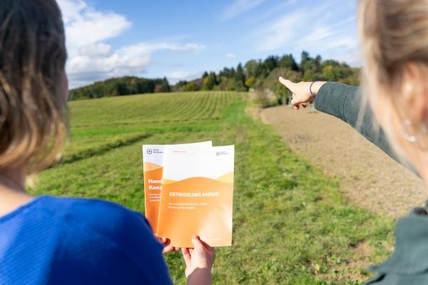 Auf dem Bild zu sehen sind zwei Frauen, die eine hält zwei orangene Broschüren ins Bild. Dahinter ist das Baugebiert Hafner zu sehen - aktuell noch unbebaut, Wiese und Acker.