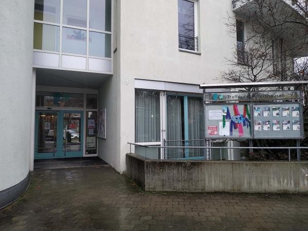 Zu sehen ist eine Gebäudefassade mit mehreren Fenstern und eine Eingangstür. Zudem eine Möglichkeit, aktuelle Informationen an eine Außenwand zu hängen. Darauf angebracht sind bereits mehrere Zettel.