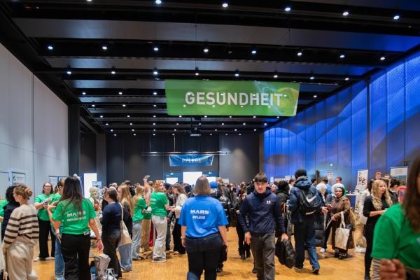 Menschen stehen verteilt in einer Halle. Von der Decke hängt ein grünes Banner mit der weißen Schrift "Gesundheit"