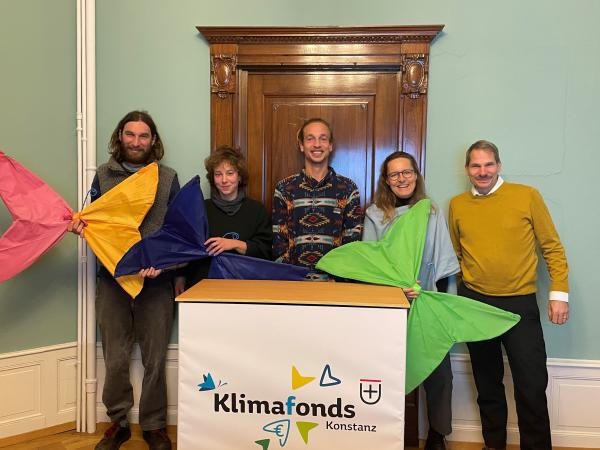 Fünf Personen stehen nebeneinander vor einer braunen Holztür. Drei halten bunte Stoff-Schmetterlinge in den Händen. Vor den Personen steht ein Counter mit dem Aufdruck "Klimafonds Konstanz"
