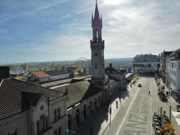 Auf dem Bild zu sehen ist der Bahnhofplatz von oben. Das Bahnhofsgebäude auf der linken Bildseite, das Lago im Hintergrund und Blauer Himmel.