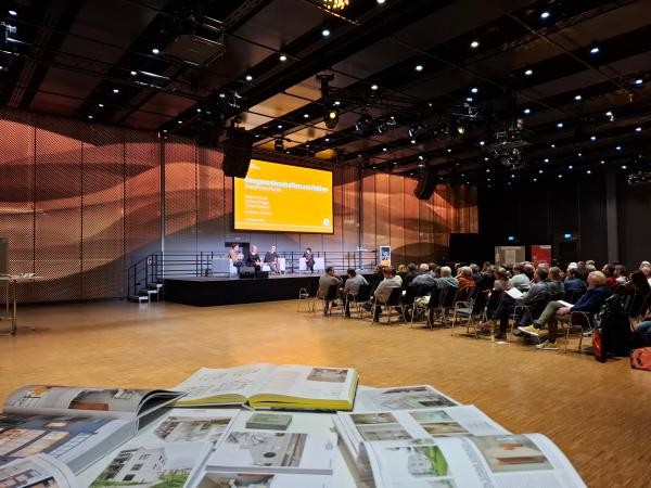 Copyright Stadt Konstanz Große Halle mit Publikum und Bühne im Hintergrund, im Vordergrund Tisch mit Informationsbroschüren