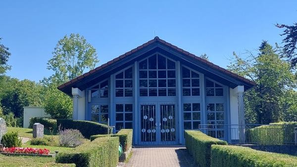 Bild eines Gebäudes mit Glasfenstern, davor Begrünung und ein Weg der in den Eingang des Gebäudes führt. Der Himmel ist blau.