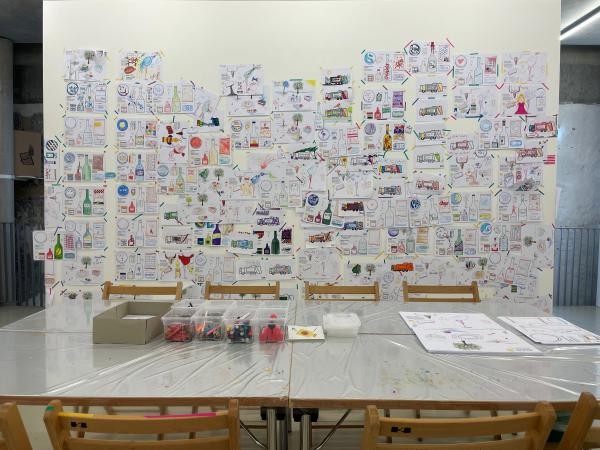 Ein großer Tisch steht vor einer weißen Wand, an der viele Kinderzeichnungen hängen.