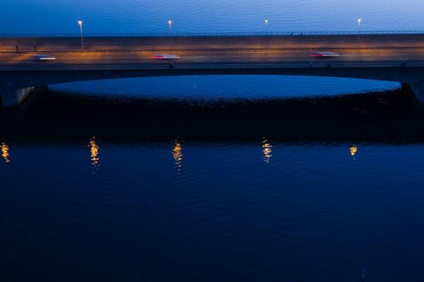 Abendaufnahme der Europabrücke in Konstanz mit sanft leuchtender Straßenbeleuchtung. Die warmen Lichtpunkte spiegeln sich leicht im tiefblauen Wasser des Rheins, während Autos die Brücke überqueren.
