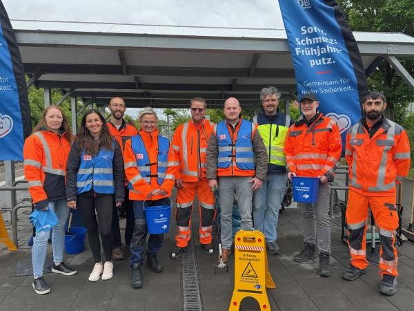 Neun Personen in orangefarbenen und blauen DB-Arbeitsschutzwesten stehen gemeinsam am Haltepunkt Wollmatingen. Sie halten Putzeimer und Reinigungsgeräte. Im Hintergrund sind Fahnen mit der Aufschrift „Sorry, Schmutz: Frühjahrsputz – Gemeinsam für mehr Sauberkeit" zu sehen.
