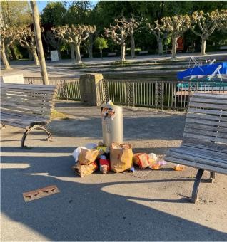 Überquellender Mülleimer zwischen zwei Parkbänken; drumherum türmt sich weiterer Müll