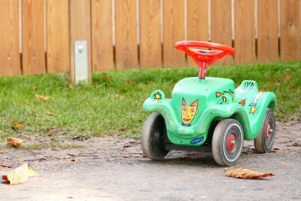 Ein grünes Spielzeugauto mit rotem Lenkrad und schwarzen Reifen zum Draufsitzen für ein Kind vor einem Holzzaun mit etwas Gras.