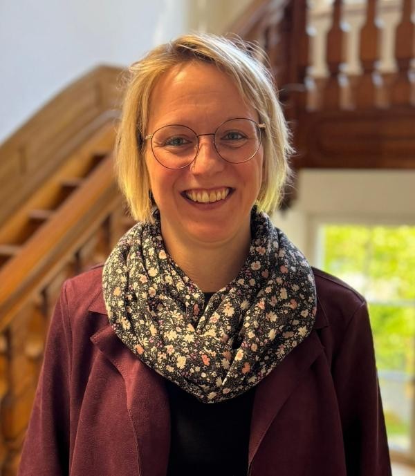 Foto einer blonden Frau mit kinnlangen Haaren, Brille und Schal. Im Hintergrund eine Holztreppe