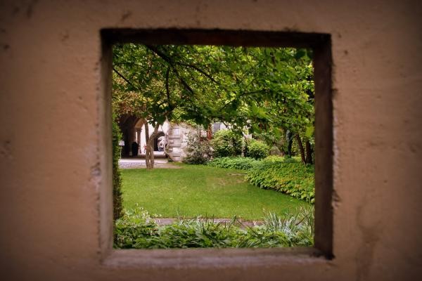 Quadratisches Loch in einer braunen Wand, durch das ein grüner Innenhof mit Bäumen zu sehen ist