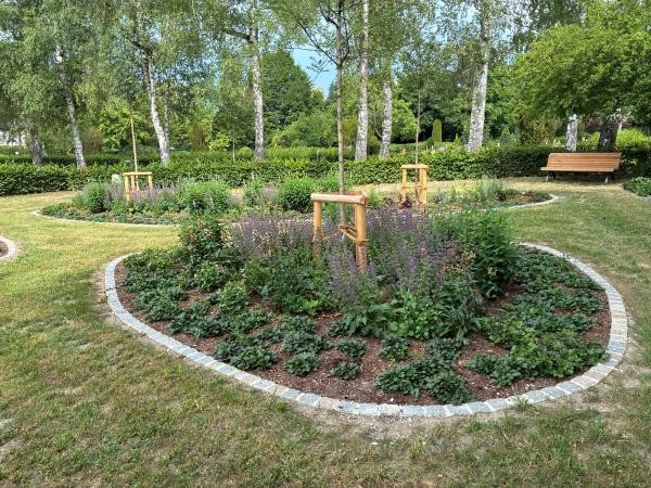 Baumgrabfeld in einem gepflegten Park: mehrere runde, mit Steinen eingefasste Pflanzbeete umgeben die Ruheplätze, dazwischen Rasenflächen. Im Beet wachsen blühende Stauden, unter hohen Bäumen stehen kleine hölzerne Gestelle, und rechts ist eine Bank zu sehen.