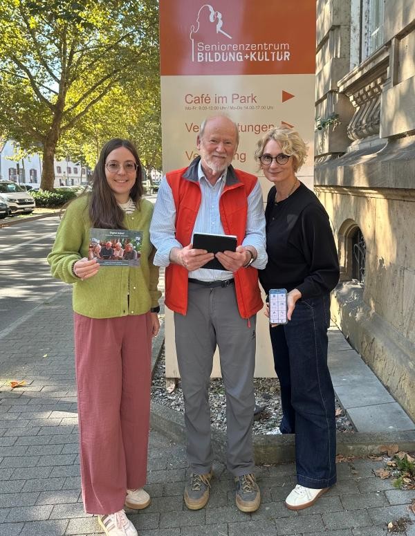 Copyright Stadt Konstanz zwei Frauen und ein Mann in der Mitte stehen vor einer Stehle mit der Aufschrift Seniorenzentrum Bildung +Kultur. Im Hintergrund ist ein Baum zu sehen