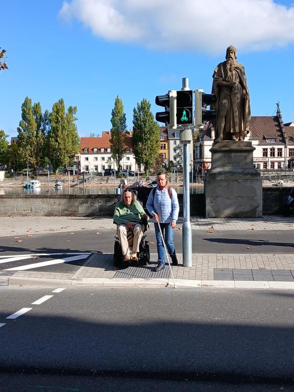 Ein Mann im Rollstuhl und eine Frau mit Blindenstock warten an einer grünen Fußgängerampel am Rheinsteig vor der Berthold-I.-Statue. Im Hintergrund sind der Hafen mit Booten sowie historische Gebäude der Konstanzer Altstadt zu sehen.