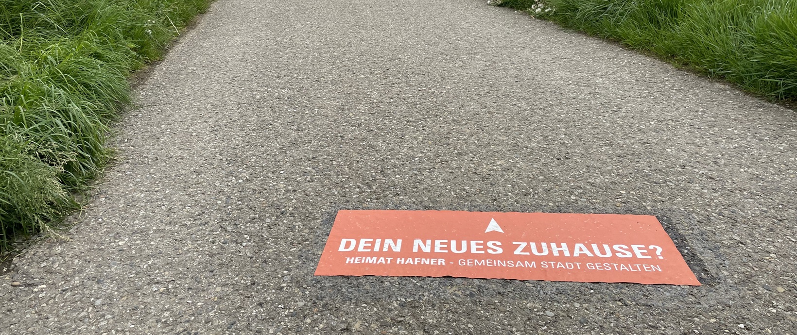 Heimat Hafner Asphaltierter Weg mit Aufkleber mit der Aufschrift "Dein neues Zuhaue? Heimat Hafner - Gemeinsam Stadt gestalten"