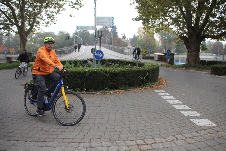 Das Foto zeigt bei grauem Wetter einen älteren Mann auf einem Elektro-Fahrrad. Er fährt vor dem Hintergrund einer Brücke um einen kleinen Kreisel, trägt eine grelle orange Jacke und einen Helm, sowie eine Schutzbrille und einen Schnurrbart.
