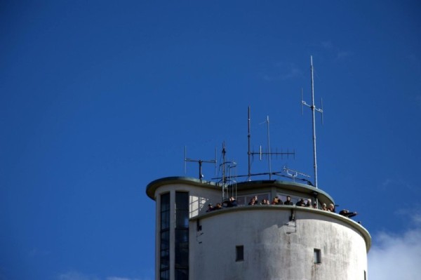 Blick von unten auf den runden Turm der Jugendherberge Konstanz-Allmannsdorf vor strahlend blauem Himmel. Auf der umlaufenden Dachterrasse stehen mehrere Menschen. Auf dem Dach sind verschiedene Antennen montiert.