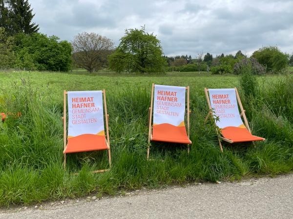 Drei Klappstühle in orange weiß auf grüner Wiese, dahinter Landschaft und Himmel.