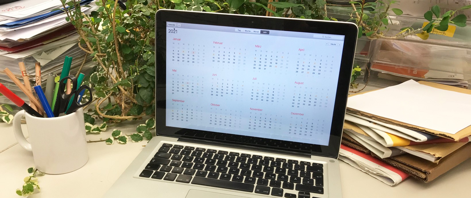 Laptop mit Kalenderansicht auf Schreibtisch, im Hintergrund Pflanzen, Zeitschriften und Schreibutensilien