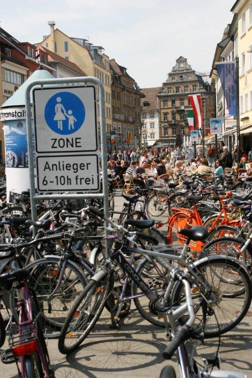 Schild „ZONE“ mit Fußgänger- und Radfahrer-Symbol, darunter „Anlieger 6–10h frei“ an einer stark frequentierten Innenstadtstraße. Vor und neben dem Schild sind zahlreiche Fahrräder dicht an dicht abgestellt; im Hintergrund sitzen Menschen vor Restaurants.