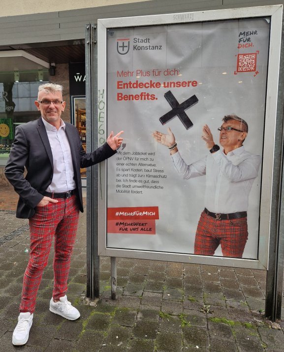 Ein Mann steht neben einem Plakat und zeigt darauf. Auf dem Plakat ist er selbst abgebildet.