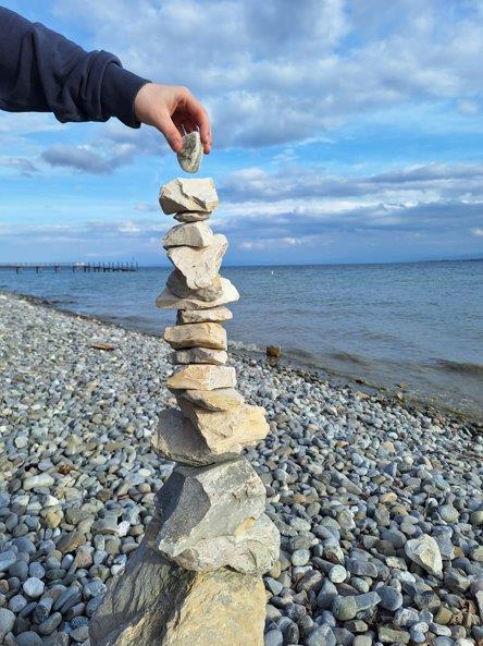 Ein Turm aus steinen am Bodenseeufer, eine Hand legt noch einen weitern Steinoben drauf