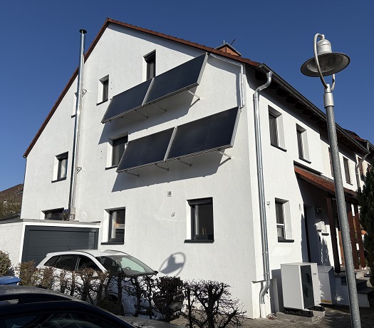 Foto eines Gebäudes mit Solarthermiekollektoren an der Fassade und einer Wärmepumpe davor.