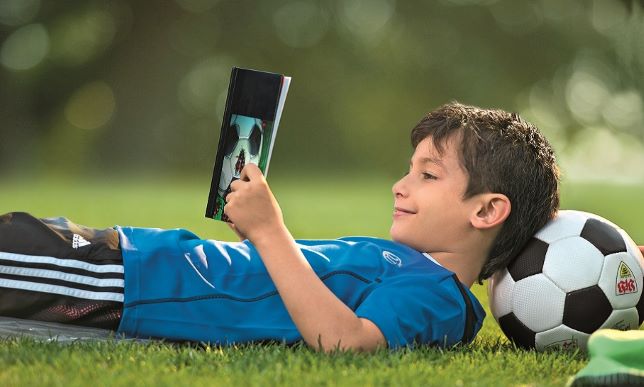 Ein Junge liegt auf dem Rücken auf dem Rasen. Er trägt ein blaues Sportshirt, hat den Kopf auf einem Fußball abgestützt und liest ein Buch.