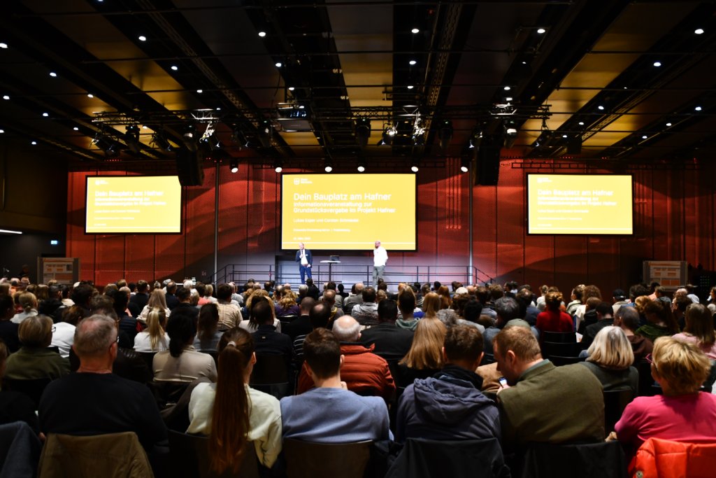 Copyright Stadt Konstanz sitzendes Publikum mit Blick auf die Bühne und zwei Personen auf der Bühne dahinter eine Präsentation
