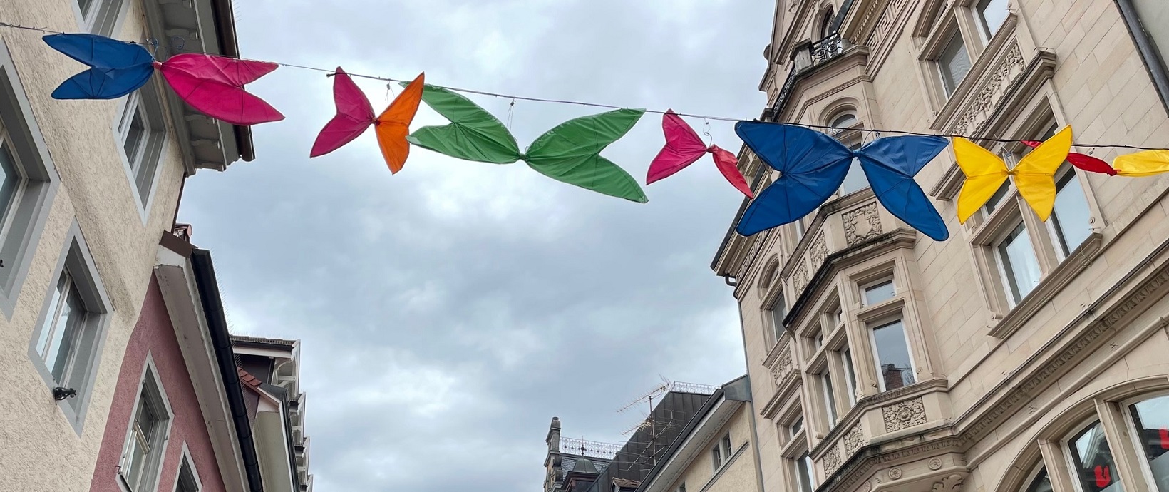 Zwischen Gebäudefassaden spannt sich eine Leine mit bunten Stoff-Schmetterlingen