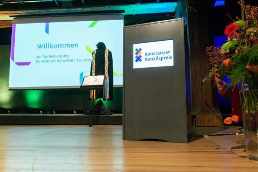 Blick auf die Bühne, man sieht einen Blumenstrauß und ein Rednerpult im Vordergrund, im Hintergrund eine Projektion auf der "Willkommen zur Verleihung des Konstanzer Konzilspreises 2019" steht.on