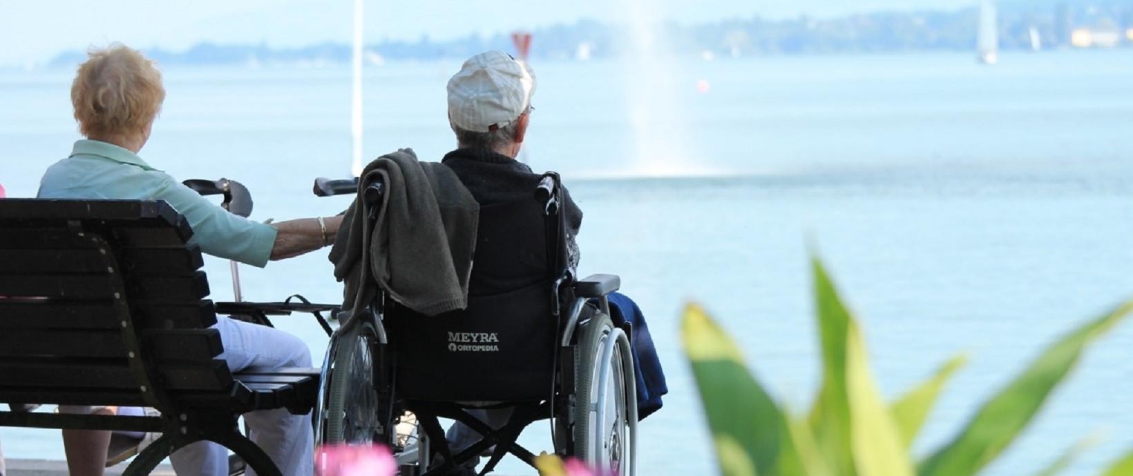 Eine Frau auf einer Bank und ein Mann im Rollstuhl schauen auf den See